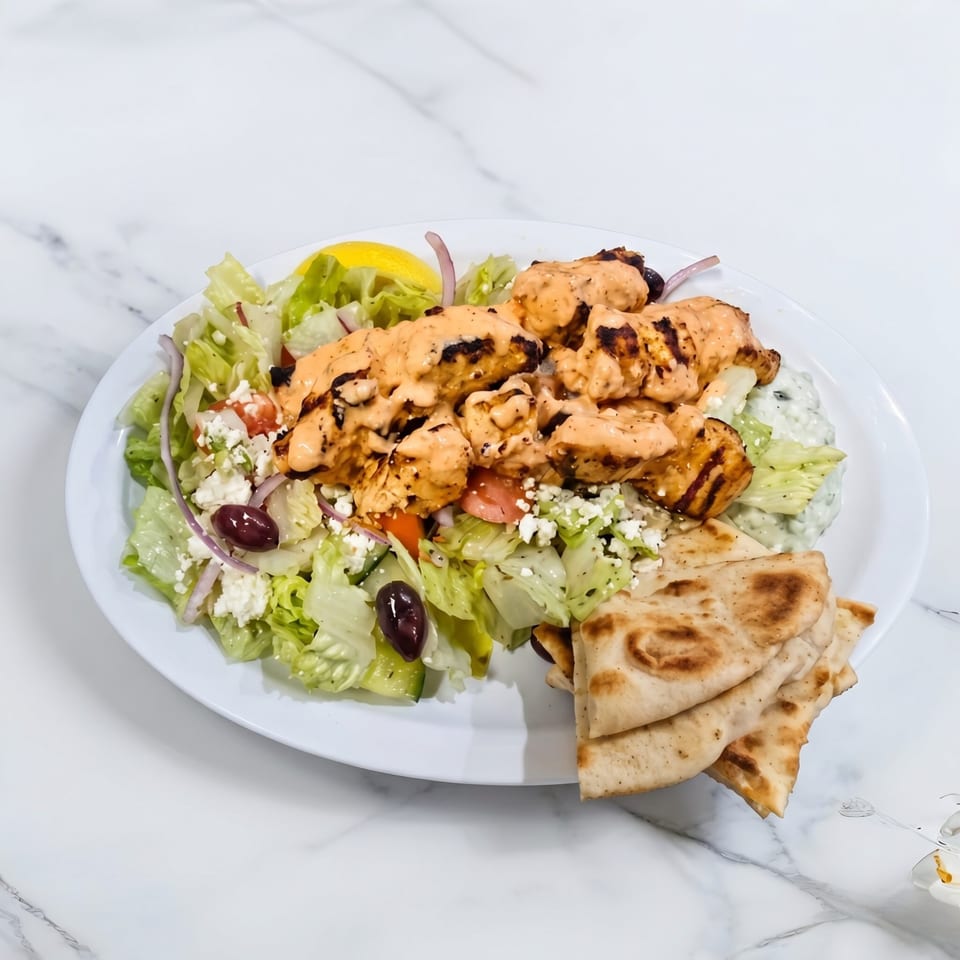 Grilled chicken skewers with creamy sauce, Greek salad, tzatziki, and pita bread on a white plate.