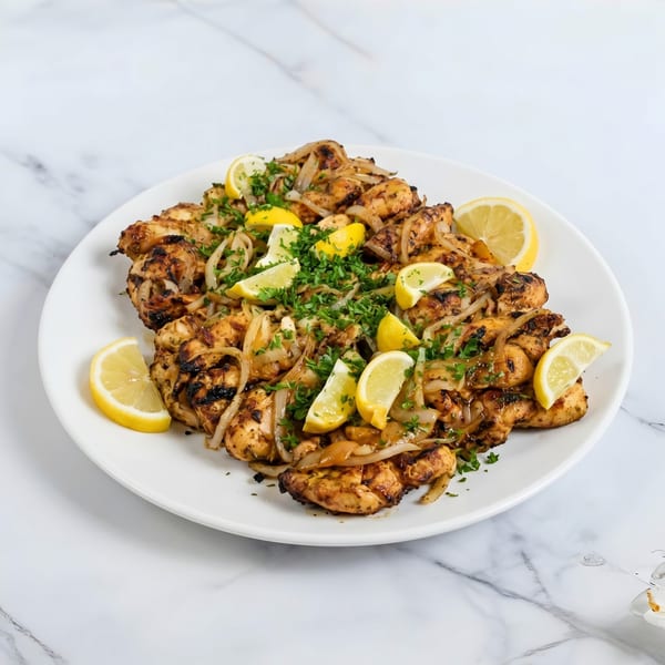 Grilled chicken pieces with sautéed onions, fresh parsley, and lemon wedges on a white plate.